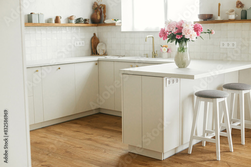 Modern minimal kitchen interior. Beautiful peonies in vase on granite countertop island on background of stylish white kitchen with wooden shelves and appliances in new house