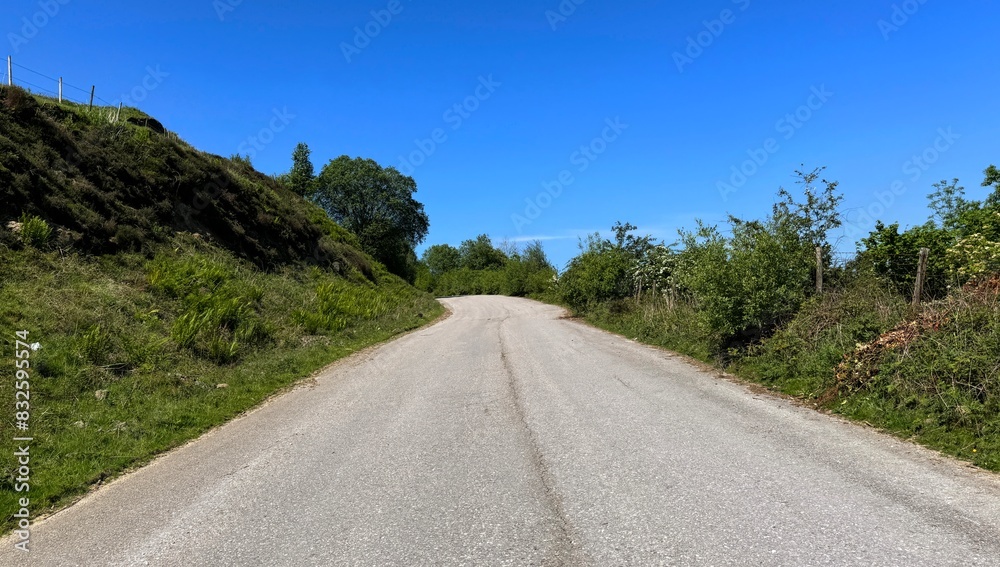 Hutch Bank Road stretches ahead, flanked by greenery under a clear blue sky, the asphalt road curves upwards and out of view in, Haslingden, Lancashire, UK