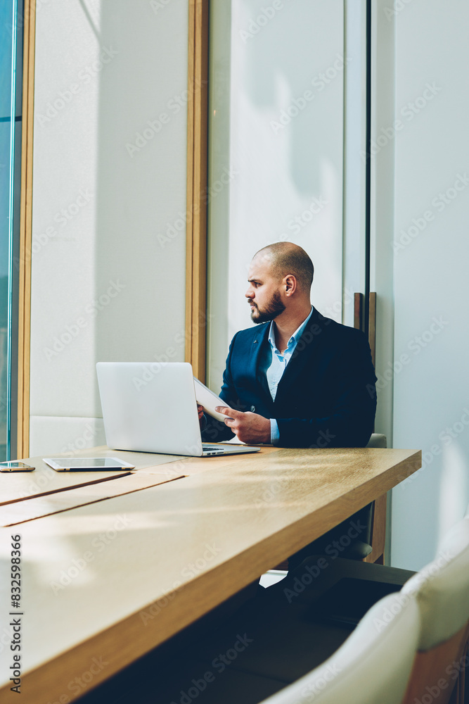 Serious businessman with accounting reports in hands sitting at laptop computer in own office while looking out of window.Confident corporate director working at modern netbook at meeting table Serious businessman with accounting reports in hands sitting at laptop computer in own office while looking out of window.Confident corporate director working at modern netbook at meeting table