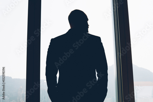 Back view of male banker dressed in black elegant suit holding hands in pockets and looking out of window while thinking on new financial project after hard working day standing in office interior