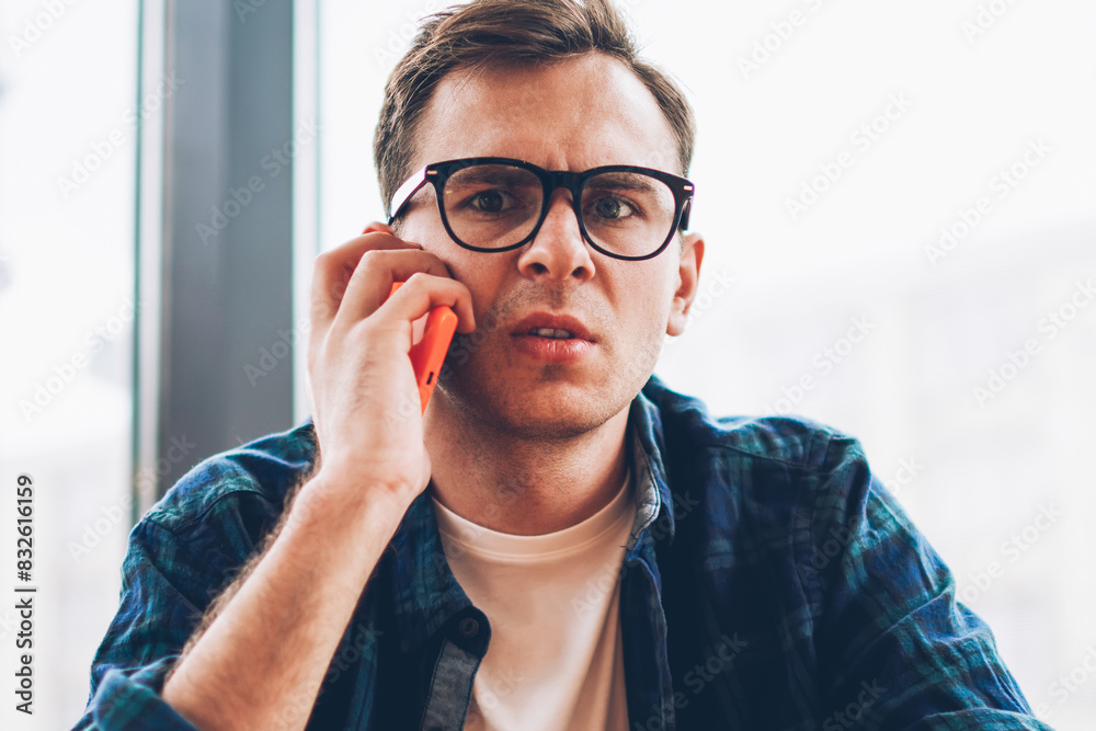 Half length portrait of pondering male student in cool spectacles communicating with friends via telephone connected to 4G.Young surprised man looking at camera while talking on cellular indoors Half length portrait of pondering male student in cool spectacles communicating with friends via telephone connected to 4G.Young surprised man looking at camera while talking on cellular indoors
