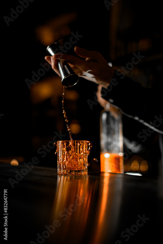 Bartender's hands pour from a jigger the last drops of alcoholic drink into a low glass