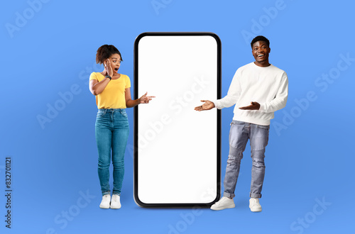 A man and a woman are standing side by side, looking at a blank screen on a phone. The man is holding the phone, while the woman is gesturing towards it with a curious expression.