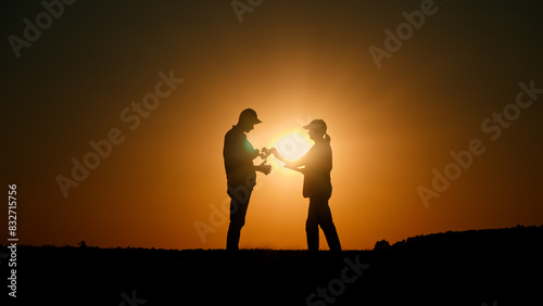 At sunset  a couple of farmers are in a field  inspecting a plant while referring to a tablet.