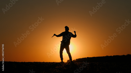 A man with a tablet in his hand dances a cheerful dance in a field at sunset