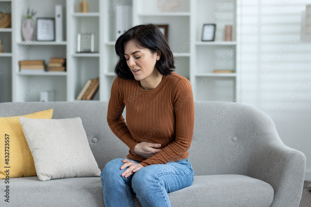 A young woman grimacing in pain is sitting on the sofa at home and holding her stomach with her hand, feeling severe pain A young woman grimacing in pain is sitting on the sofa at home and holding her stomach with her hand, feeling severe pain