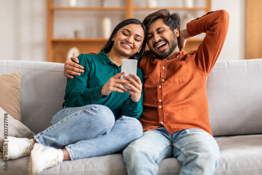 Indian man and woman are seated on a couch, both engrossed in a cell phone. They appear focused on the screen, possibly sharing content or engaging in a conversation related to the device. Indian man and woman are seated on a couch, both engrossed in a cell phone. They appear focused on the screen, possibly sharing content or engaging in a conversation related to the device.