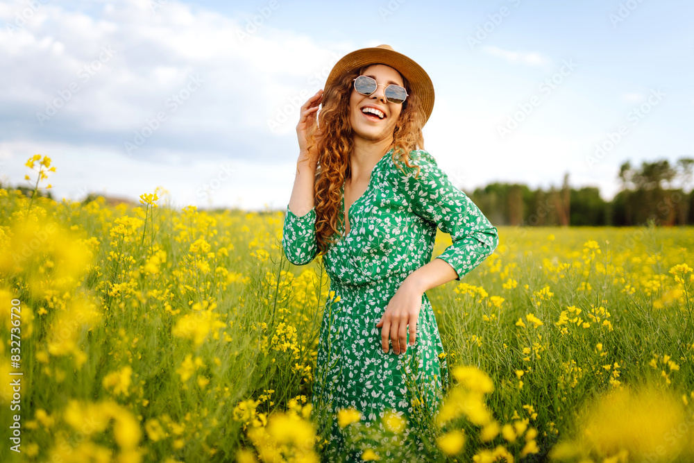 Fototapeta premium A happy woman in a hat walks through a blooming rapeseed field. Nature, rest. Summer landscape.