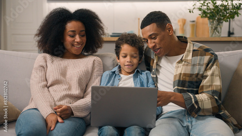 Happy African American family laughing woman mom female cheerful dad man male little boy son child kid watch movie cartoon using laptop computer pc playing game talking enjoy online shopping sofa home