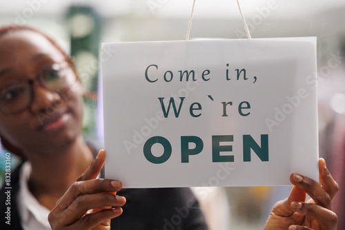 Fashion clothes shop manager holding opening signboard with come in message. Apparel store young african american woman assistant hanging sign informing clients about workday beginning
