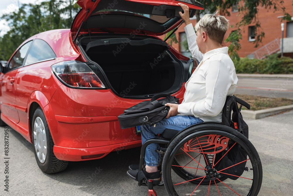 Woman with disability on wheelchair opening the trunk of her car to put the laptop before driving to work. Independency concept. Woman with disability on wheelchair opening the trunk of her car to put the laptop before driving to work. Independency concept.