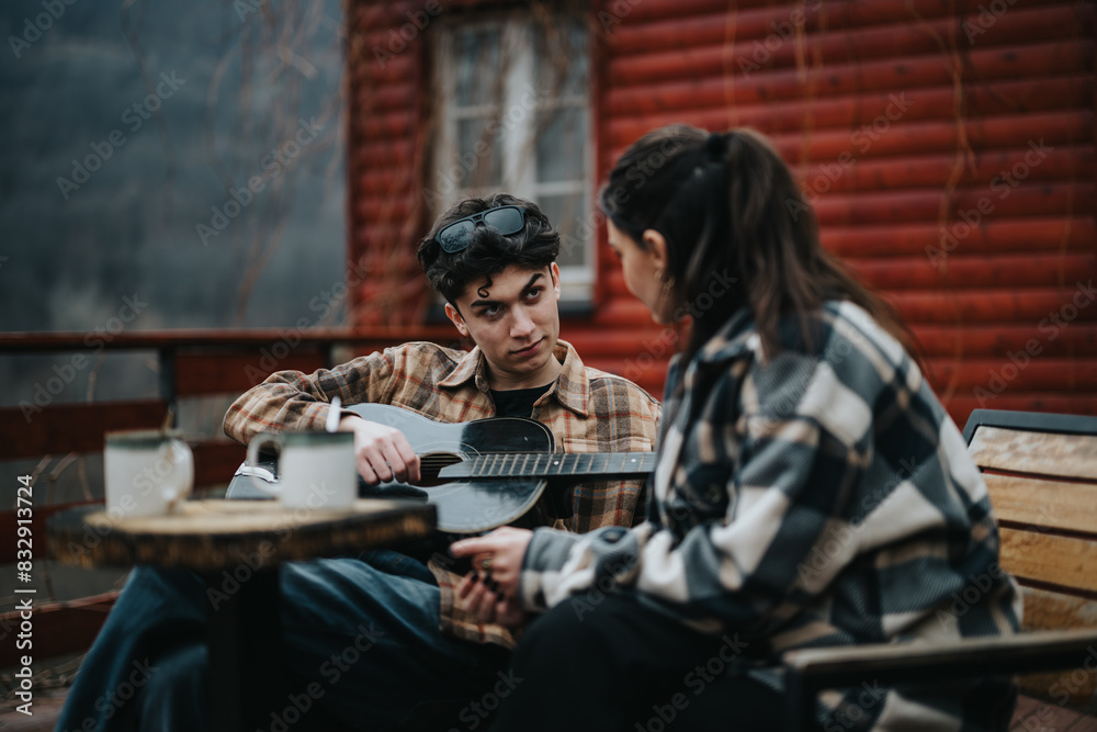 Two young adults engaged in a relaxed conversation outdoors, with a guitar and coffee on a table, by a wooden cabin.