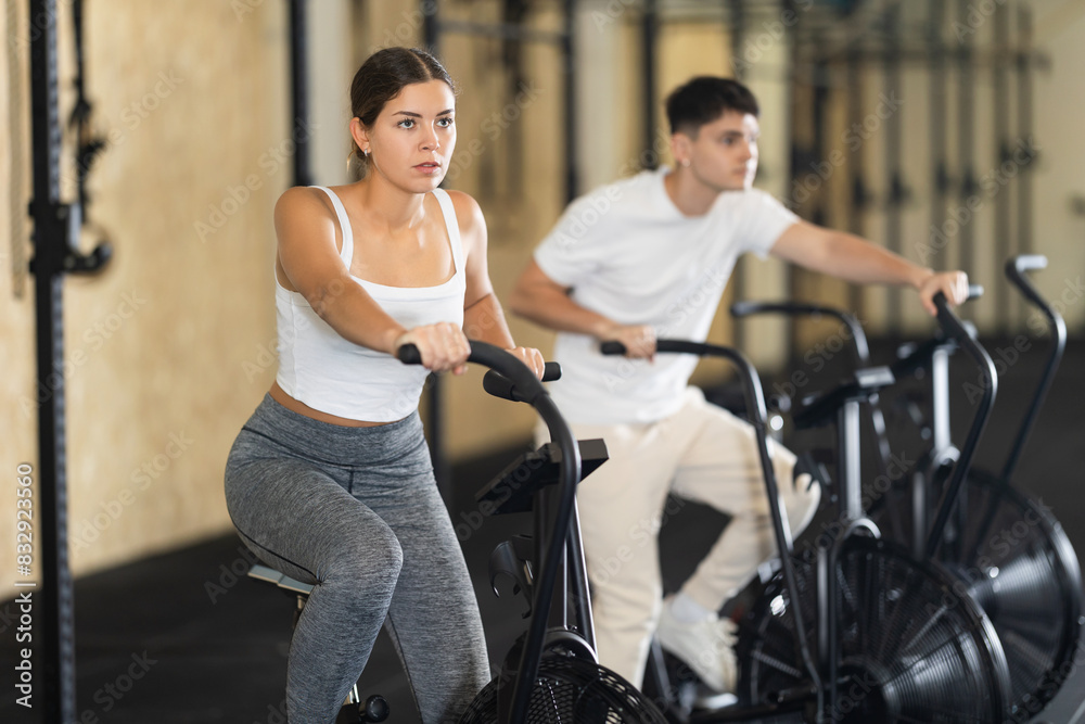 Fototapeta premium Positive girl riding exercise bike for cardio workout during cycling class at modern crossfit gym