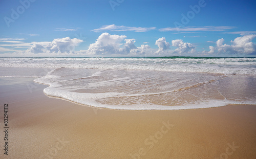 Tide coming in as waves break and roll onto seashore making foamy white patterns across the wet sandy beach, and  horizon with blue cloudy winter sky in background.