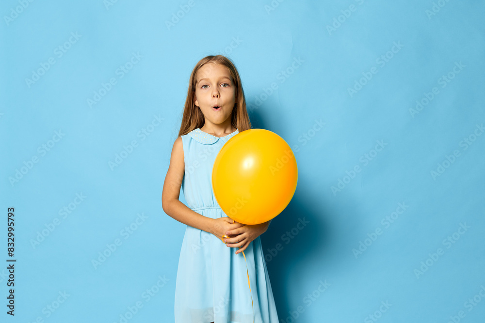 Little girl in a colorful dress joyfully holding a yellow balloon against a vibrant blue background Little girl in a colorful dress joyfully holding a yellow balloon against a vibrant blue background