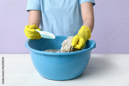 Woman in rubber gloves adding detergent to clothes in plastic basin on table against lilac background