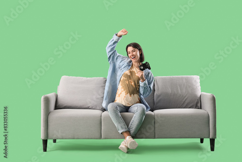 Happy young woman with gamepad sitting on sofa against green background