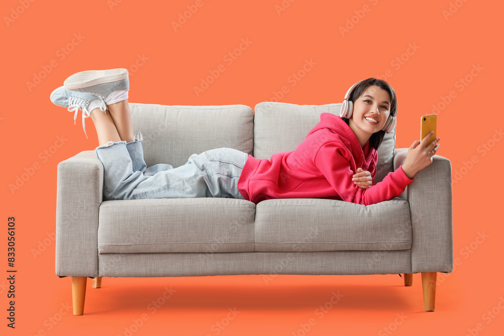 Happy young woman in headphones with mobile phone lying on sofa against orange background Happy young woman in headphones with mobile phone lying on sofa against orange background