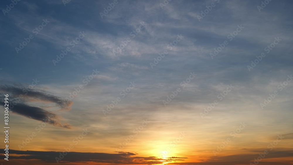 Naklejka premium A stunning sunrise with a mix of golden and orange hues at the horizon, gradually blending into the blue sky above. Wispy clouds are scattered, adding texture to the serene scene. Cloud background.