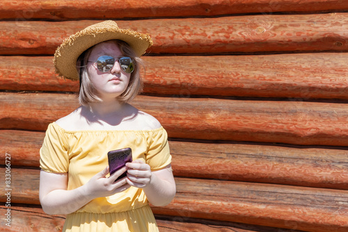 Asian teenage girl in yellow dress, hat and sunglasses makes online shopping on the phone, communicates online against the wall of a wooden village house, copy space. Summer time. Holidays