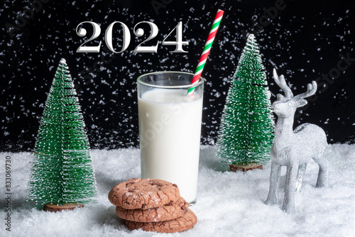 A gift for Santa. A glass of milk with a straw and cookies with chocolate chips on a snowy background among toy Christmas trees, a reindeer. New Year 2024