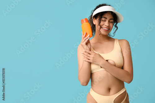 Beautiful young African-American woman in stylish beige swimsuit with bottle of sunscreen cream on blue background