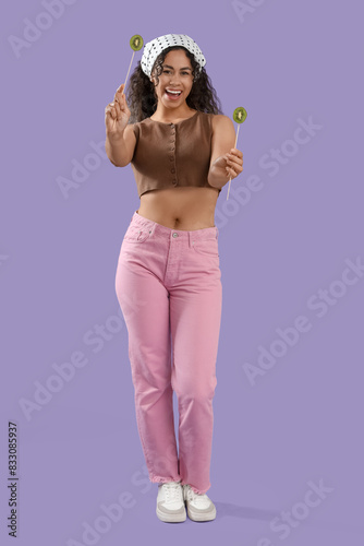 Beautiful young happy African-American woman holding sticks with kiwi on purple background © Pixel-Shot