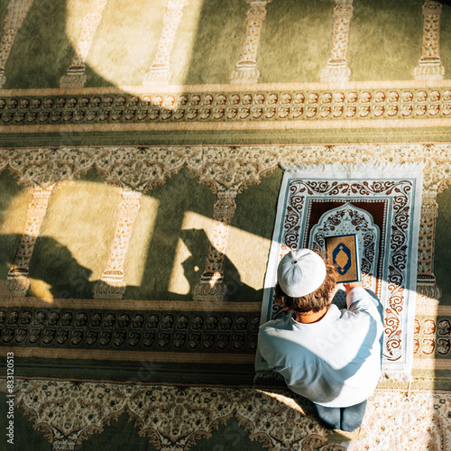 Interior of the mosque. 
Top view of a man holding and reading muslim holly book- Quran. He is standing on a green carpet in the mosque