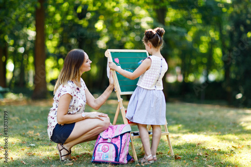 Happy little kid girl and mother by big chalk desk Preschool or schoolkid on first day of elementary class. Back to school concept. Healthy child and woman writing and painting outdoors