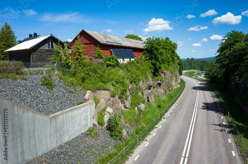 Road with a double white line  no crossing  stretching into the distance to forest  Wooden Scandinavian style farm buildings on a rock among green bushes and trees  Sunny summer day  V  rmland  Sweden
