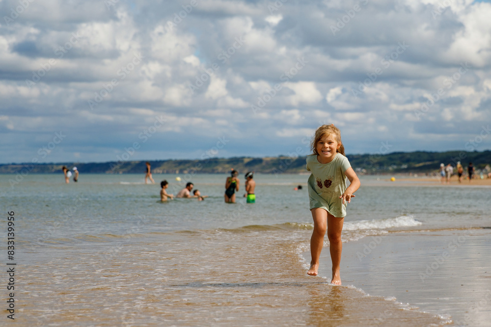 Fototapeta premium Happy preschool girl having fun on the sand beach Omaha at Atlantic coast of Normandy, France. Outdoor summer activities for kids