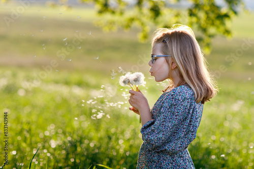 Adorable cute little girl blowing on a dandelion flower on the nature in the summer. Happy school child with eyeglasses with blowball, having fun. Bright sunset light, active kid.