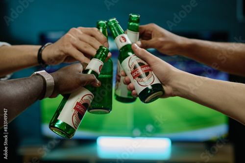 Closeup shot of hands of unrecognizable men clinking bottles with beer while watching soccer game on TV photo
