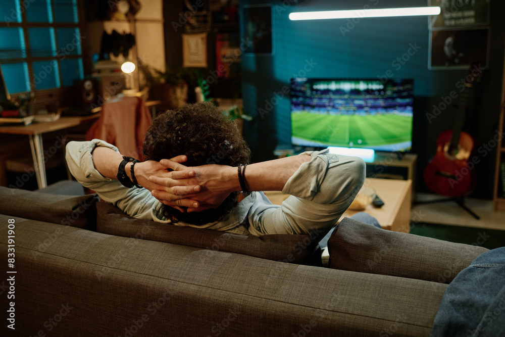 Fototapeta premium Rear view shot of unrecognizable man sitting relaxed in living room watching sports event on TV channel, copy space