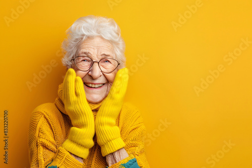 Cheerful elderly lady in a yellow sweater and colorful gloves smiles against a yellow background