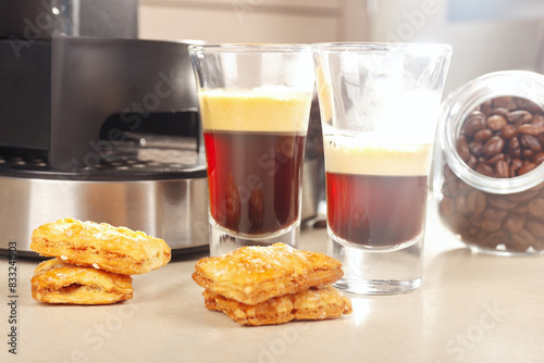 Two glass shorts of coffee near coffee maker, crackers and cans of coffee beans on light table.