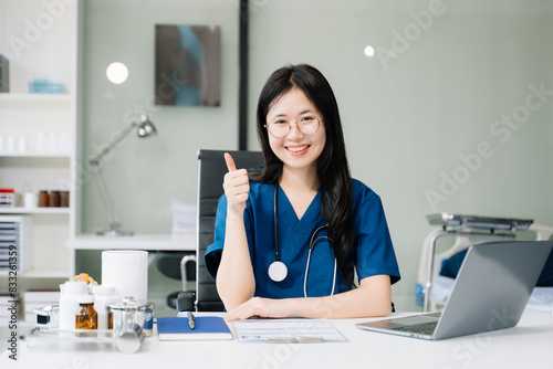 Confident young female doctor in white medical uniform sit at desk working on computer. Smiling use laptop write in medical journal