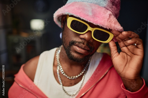 Close up portrait of Black young man wearing fuzzy bucket hat and trendy sunglasses looking at camera indoors