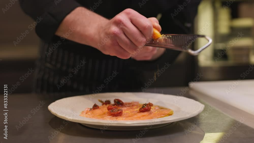 Professional chef in black working uniform grating aged cheese over dish with fish and halved cherry tomatoes. Creative gourmet adding secret ingredient to serving for food critic.