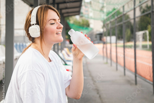 Woman in white headphones drinks water after training at the stadium, concept of outdoor sports, water balance, copy space. © sav_an_dreas