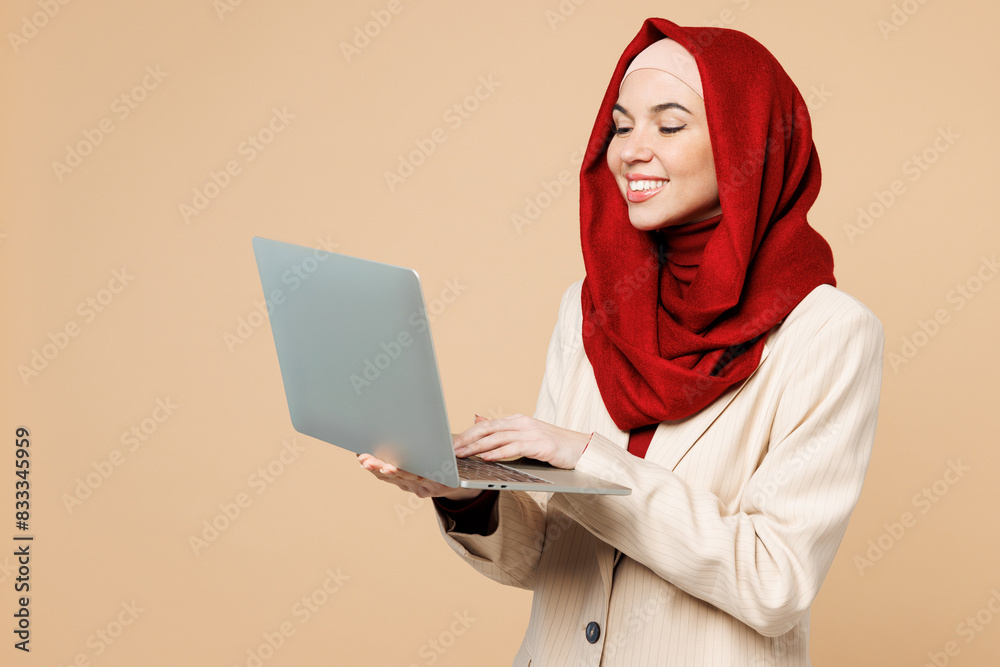 Young happy Arabian Asian Muslim IT woman wear red abaya hijab suit clothes hold use work on laptop pc computer isolated on plain beige background studio. UAE middle eastern Islam religious concept. Young happy Arabian Asian Muslim IT woman wear red abaya hijab suit clothes hold use work on laptop pc computer isolated on plain beige background studio. UAE middle eastern Islam religious concept.