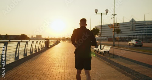 Jogging fitness workout in the city at sunset. Young athletic man training on embankment in golden light. Active healthy recreation, energy and power concept. Slow motion shot.