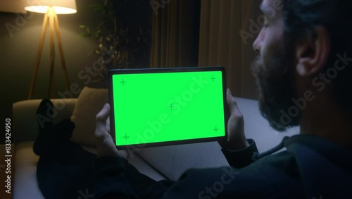 View from shoulder of man lying on couch using tablet with chroma key green screen. Man watching series or video at home at night.
