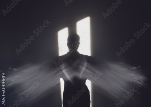 An angel with wings standing in the darkness in front of a door of light. 