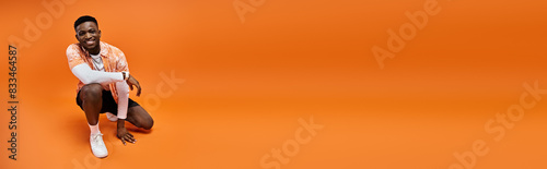 Fotografía Fashionable African American man in stylish attire crouching on vibrant orange background