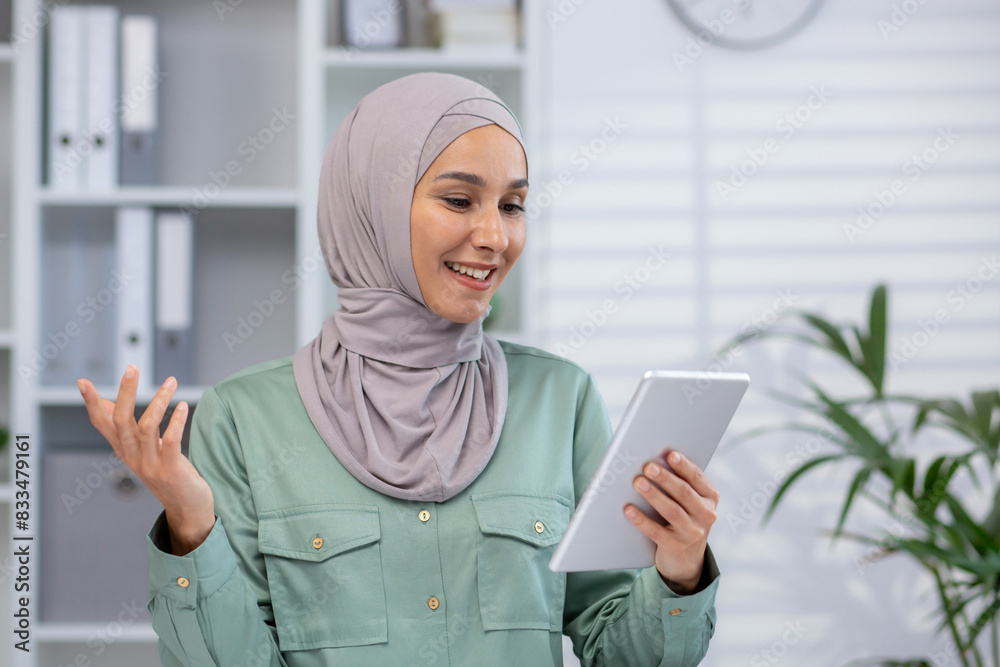 Smiling woman wearing hijab, using tablet in modern office with bookshelf and plants in the background. Smiling woman wearing hijab, using tablet in modern office with bookshelf and plants in the background.