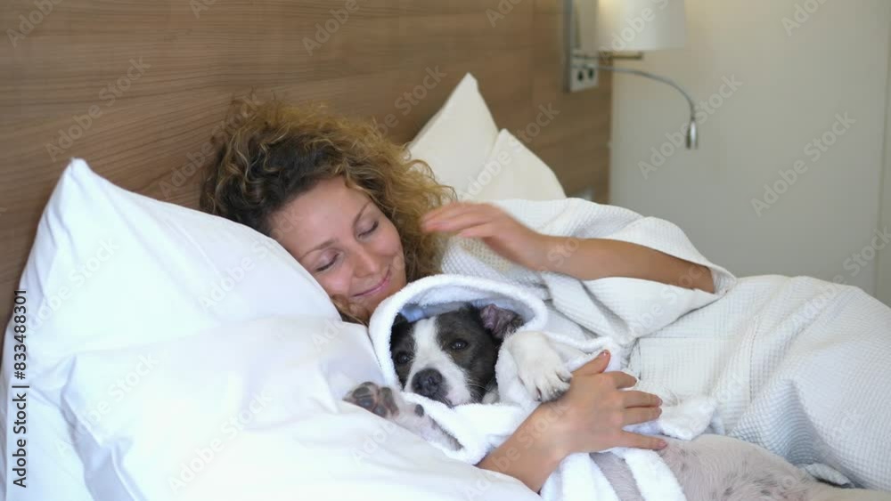 In large soft bed, woman and dog lie in white cozy bathrobes. Woman takes dog paw and waves it in friendly manner. Together in warm bathrobes, owner and dog create atmosphere of peace and friendship.