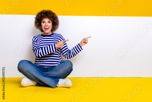 Full length photo of impressed woman dressed striped shirt sit near placard indicationg empty space isolated on yellow color background