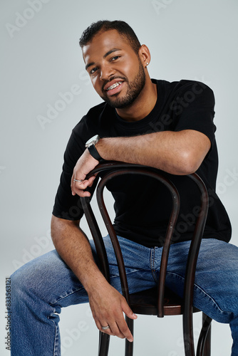Stylish African American man sitting gracefully on a wooden chair against a vibrant backdrop.