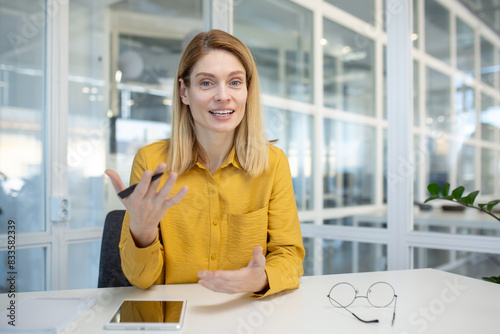 Professional woman engaging in a video call in a modern office setting, looking directly at the camera while speaking.
